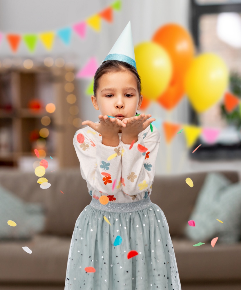 birthday, childhood and people concept - little girl in party hat blowing to confetti over decorated living room background. girl in birthday party hat blowing to confetti