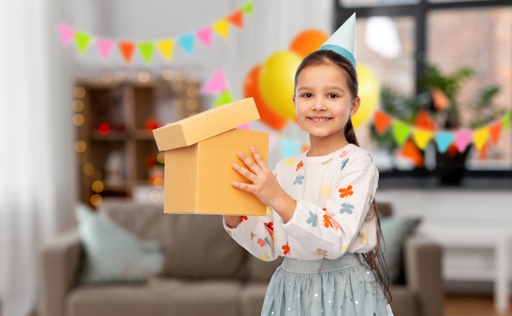 birthday, childhood and people concept - portrait of smiling little girl in party hat opening gift box over decorated living room background. smiling girl in party hat opening birthday gift