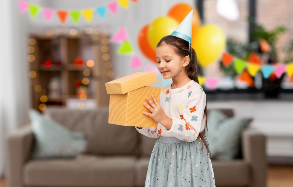 birthday, childhood and people concept - portrait of smiling little girl in party hat opening gift box over decorated living room background. smiling girl in party hat opening birthday gift