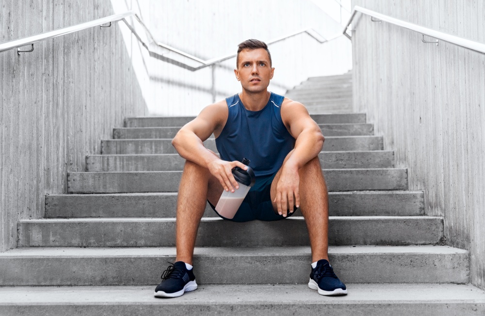 fitness, sport and people concept - tired young man with protein shake in bottle sitting on stairs. sportsman with shake in bottle sitting on stairs