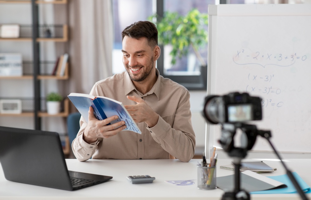 distance education, school and remote job concept - happy smiling male math teacher with camera and book having online class at home office. male math teacher having online class at home