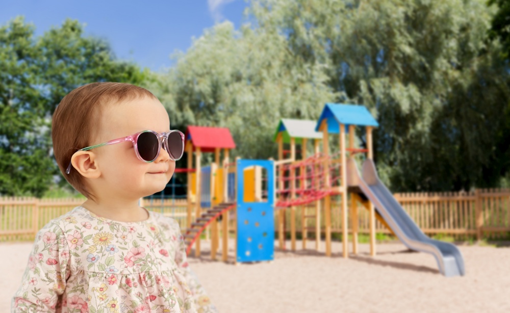 childhood, summer and people concept - happy little baby girl in sunglasses over children&rsquo;s playground background. happy baby girl in sunglasses over playground