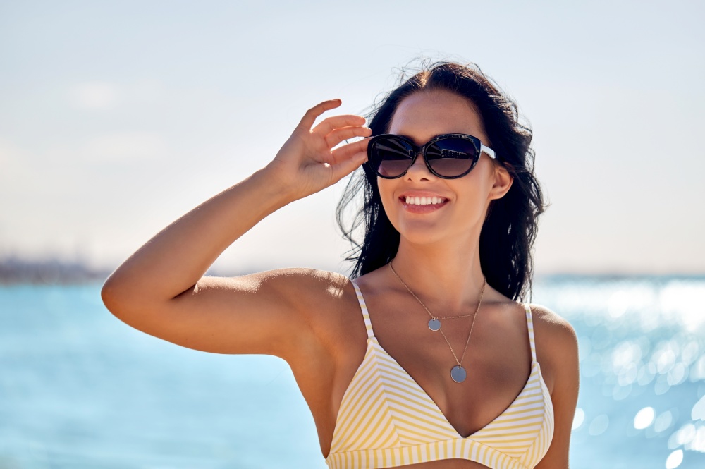 people, summer and swimwear concept - happy smiling young woman in sunglasses and bikini swimsuit on beach. smiling young woman in sunglasses on summer beach
