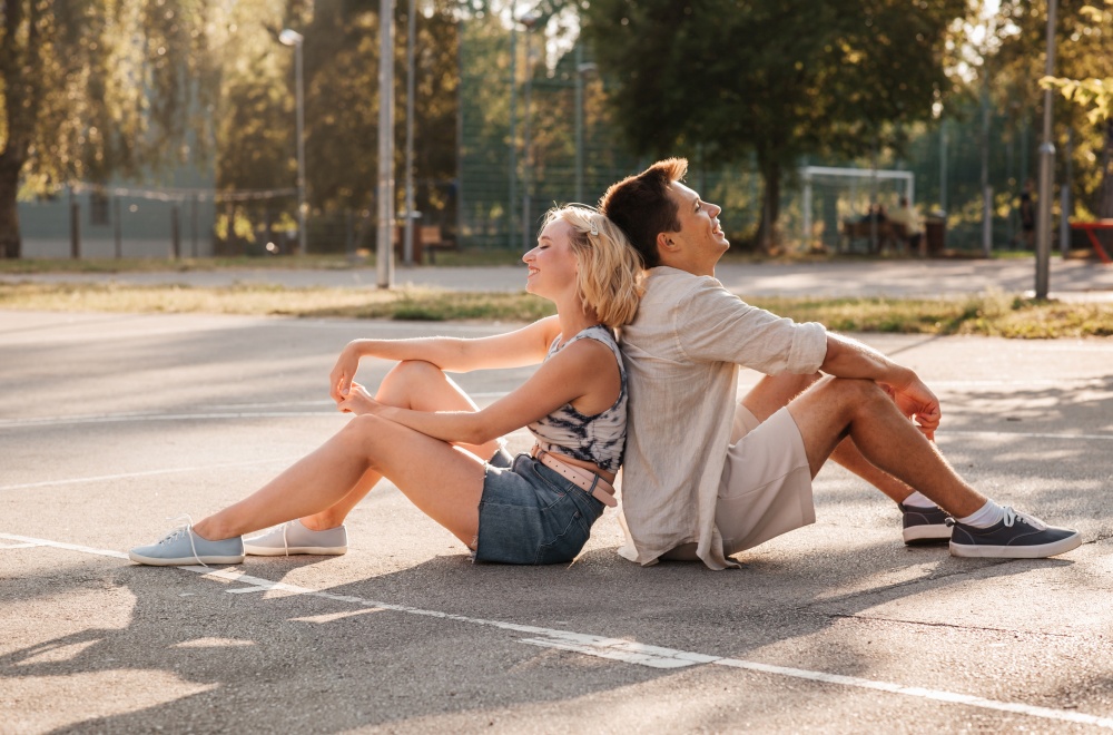 summer holidays and people concept - happy young couple sitting back to back on basketball playground. happy couple sitting on basketball playground