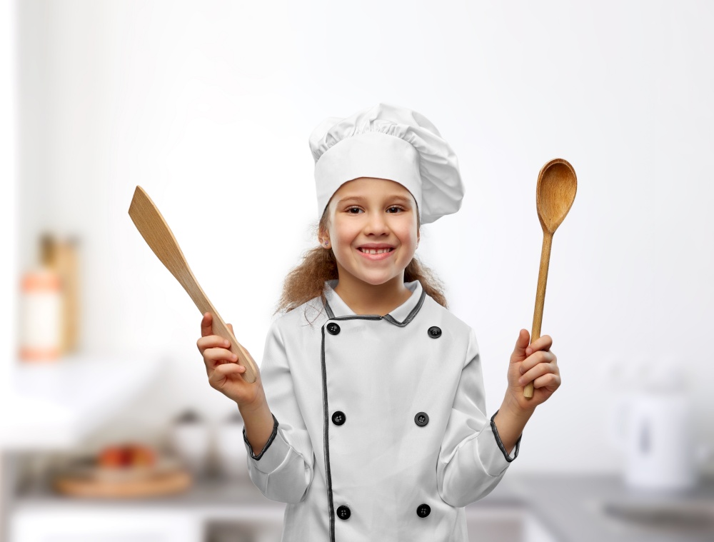 cooking, culinary and profession concept - happy smiling little girl in chef&rsquo;s toque and jacket with wooden spoon and spatula over kitchen background. happy girl in chef&rsquo;s toque with spoon and spatula