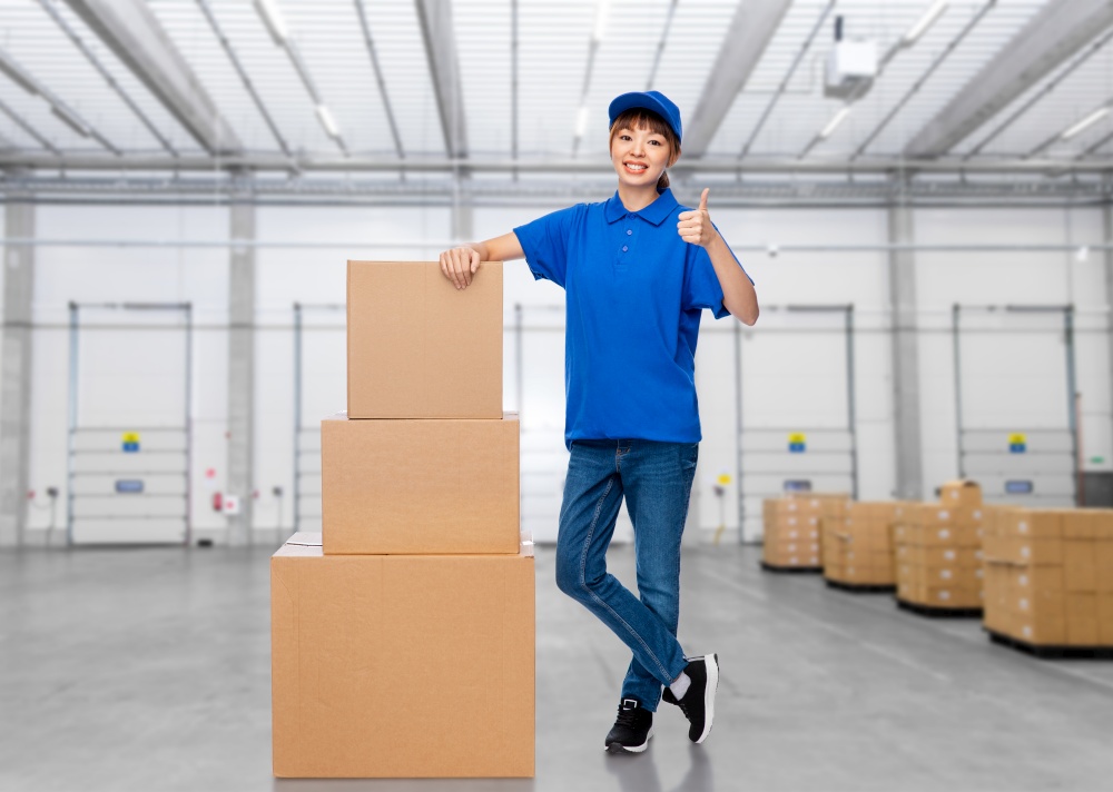 mail service and shipment concept - happy smiling delivery woman with parcel boxes in blue uniform over warehouse background. delivery woman with parcel boxes at warehouse