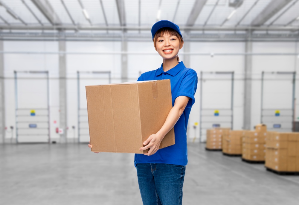 mail service and shipment concept - happy smiling delivery woman with parcel box in blue uniform over warehouse background. delivery woman with parcel box at warehouse