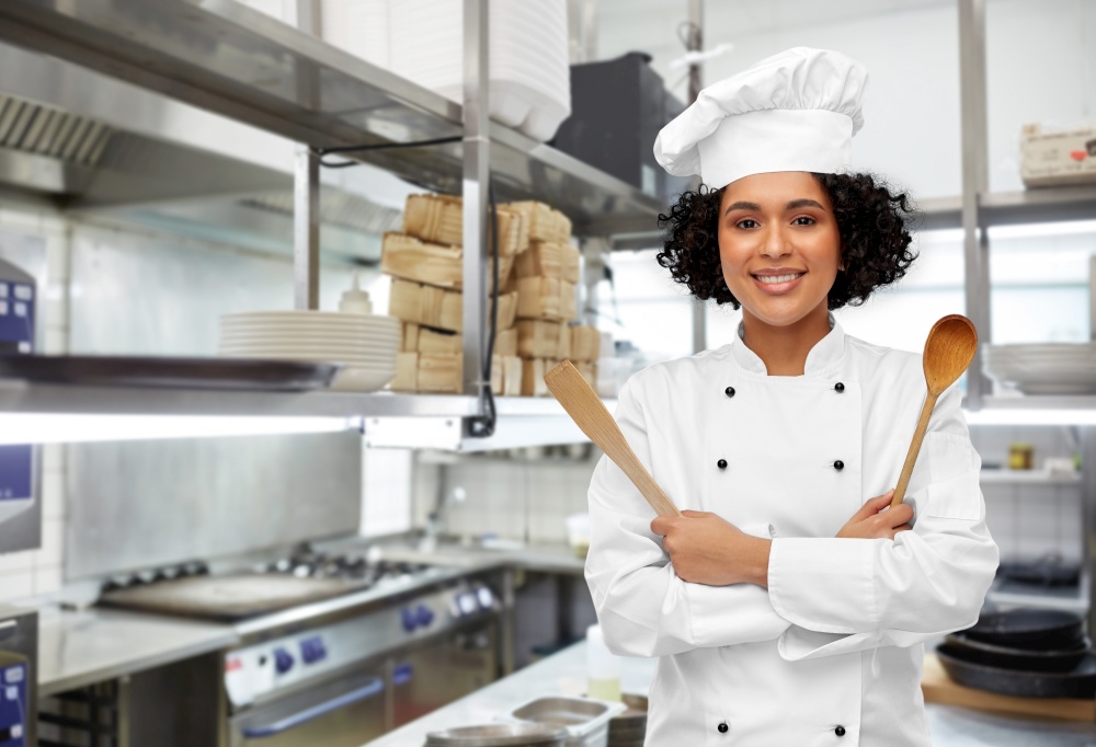 cooking, culinary and people concept - happy smiling female chef in toque with wooden spoon and spatula over restaurant kitchen background. smiling female chef with wooden spoon and spatula