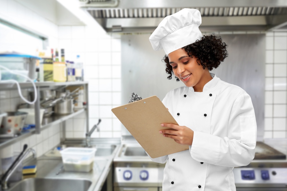 cooking, culinary and people concept - happy smiling female chef in toque and jacket with clipboard over restaurant kitchen background. smiling female chef with clipboard