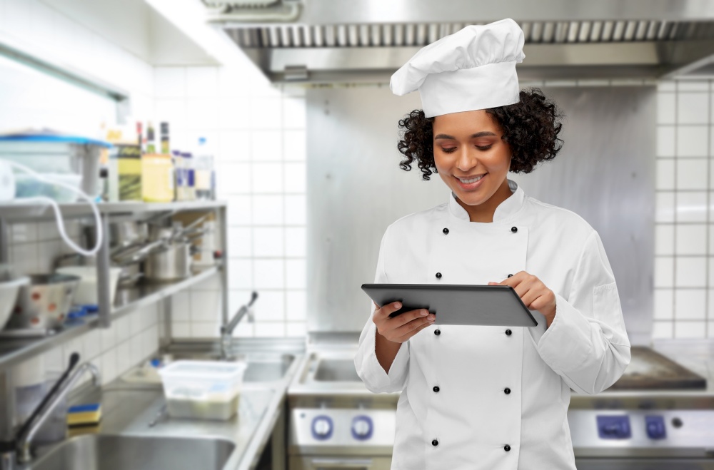cooking, culinary and people concept - happy smiling female chef in toque and jacket with tablet pc computer over restaurant kitchen background. smiling female chef with tablet computer