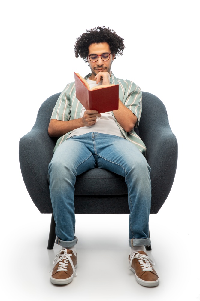 people and furniture concept - thinking young man in glasses reading book sitting in chair over white background. thinking man in glasses reading book in chair