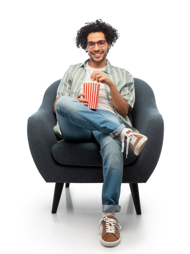 people and furniture concept - happy smiling young man in glasses with popcorn sitting in chair over white background. smiling young man with popcorn sitting in chair