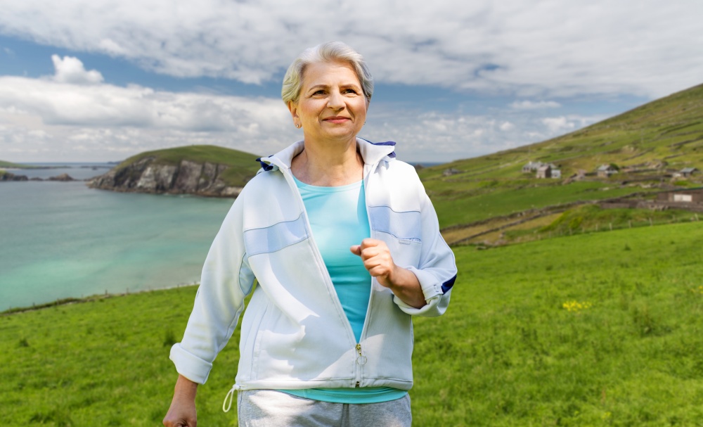 fitness, sport and healthy lifestyle concept - senior woman running over atlantic ocean coast in ireland background. senior woman running along summer park