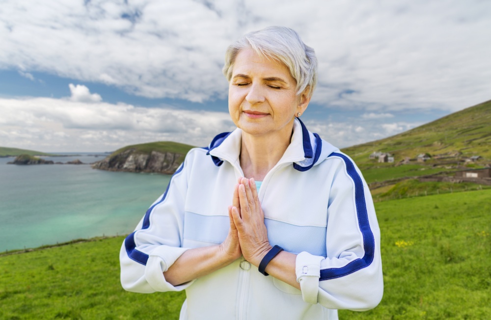 meditation, mindfulness and healthy lifestyle concept - happy smiling senior woman doing yoga outdoors over atlantic ocean coast in ireland background. happy senior woman doing yoga outdoors