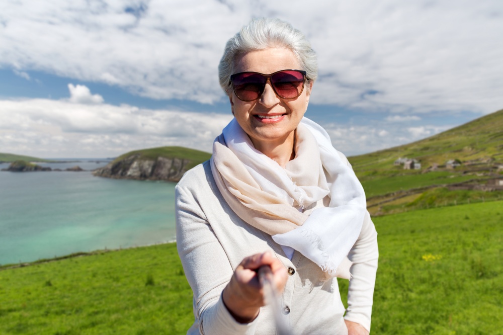 travel, tourism and vacation concept - happy smiling senior woman in sunglasses taking picture by selfie stick over atlantic ocean coast in ireland background. old woman taking picture by selfie stick on beach