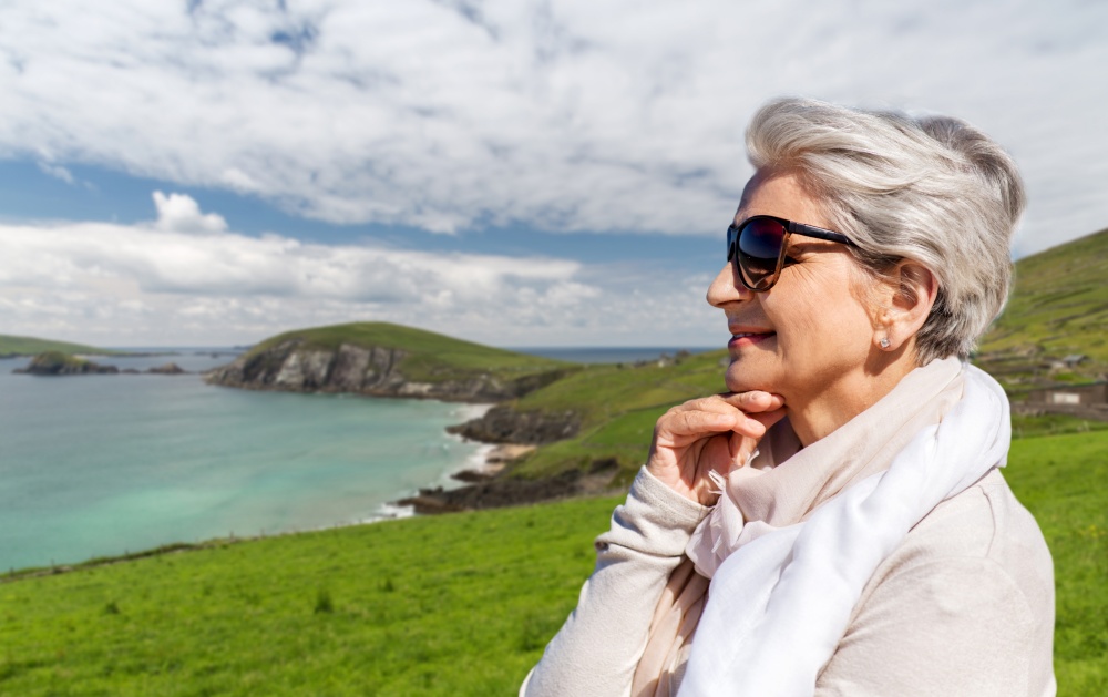 travel, tourism and vacation concept - portrait of happy senior woman in sunglasses and scarf over atlantic ocean coast in ireland background. portrait of senior woman in sunglasses on beach