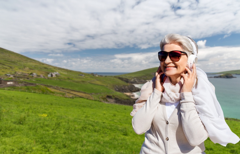 travel, tourism and vacation concept - senior woman in headphones and sunglasses listening to music over atlantic ocean coast in ireland background. old woman in headphones listens to music on beach