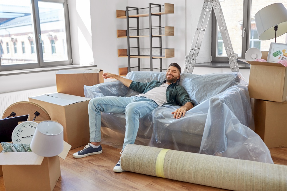 moving, people and real estate concept - happy smiling man with boxes resting on sofa covered with plastic sheeting at new home. happy man with boxes moving to new home