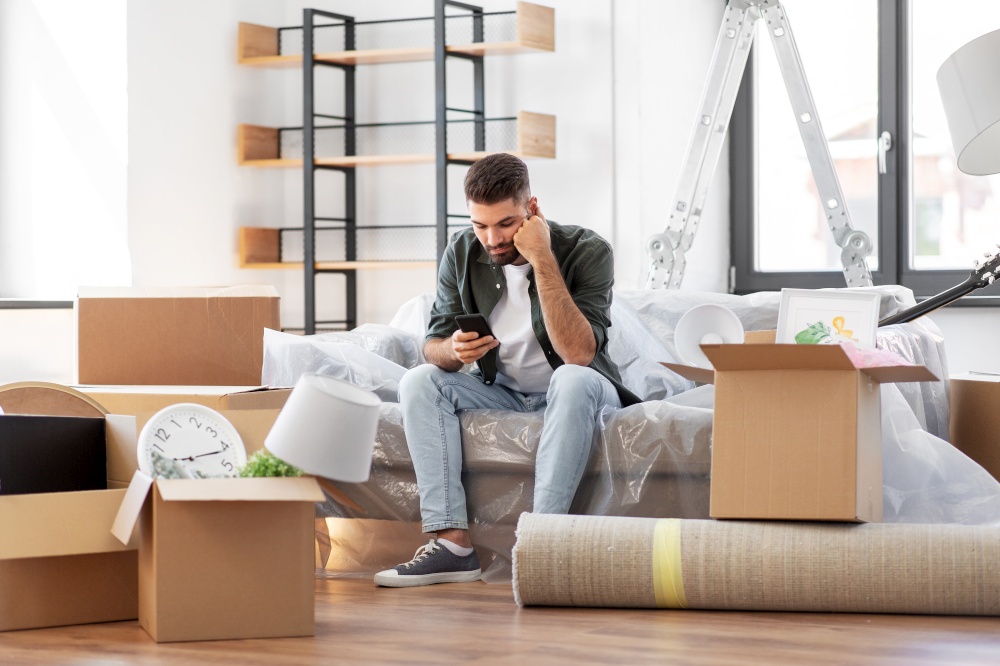 moving, eviction and real estate concept - sad man with smartphone and boxes at new home. sad man with smartphone and boxes moving home