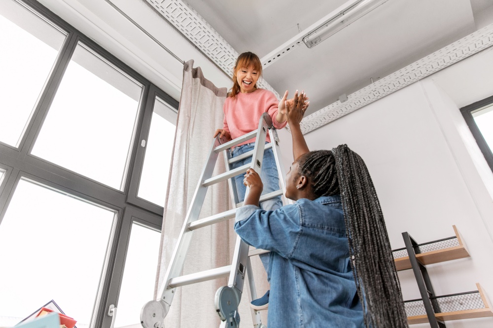 improvement and renovation concept - happy smiling women with ladder hanging curtains at home and making high five gesture. women hanging curtain at home and making high five