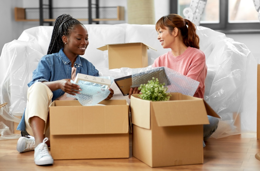 moving, people and real estate concept - women unpacking boxes at new home or packing stuff into bubble wrap. women unpacking boxes and moving to new home