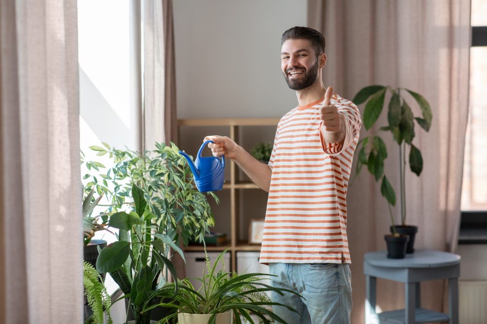 people, nature and plants care concept - happy smiling man watering flowers and showing thumbs up gesture at home. smiling man watering flowers and showing thumbs up