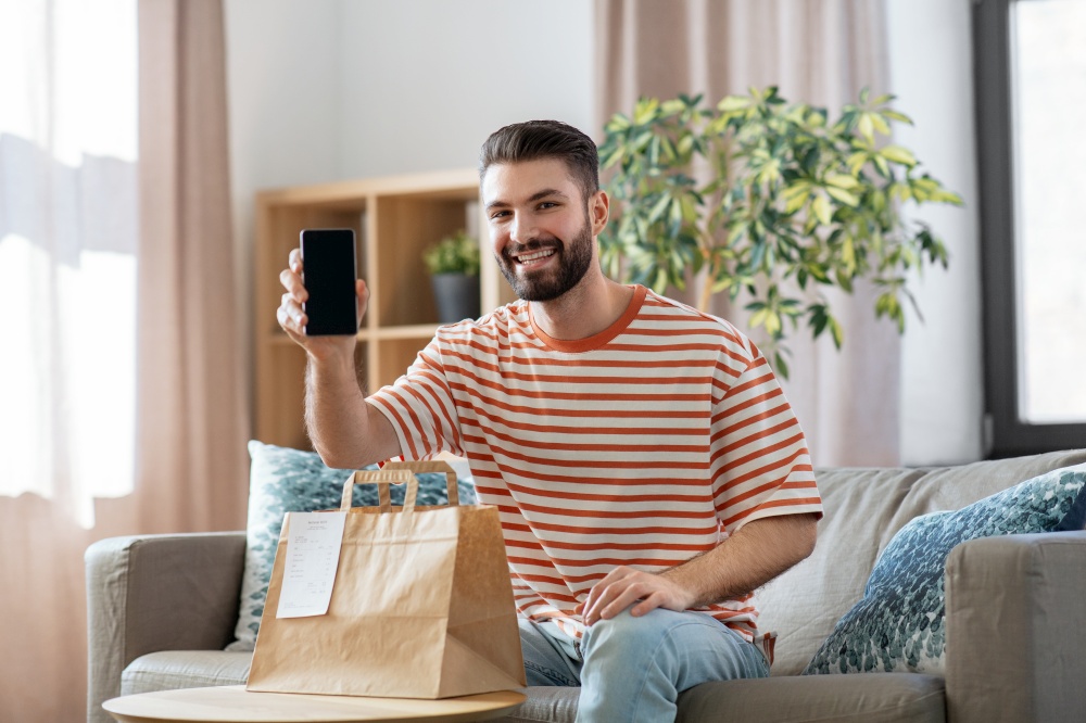technology, delivery and people concept - smiling man with smartphone and takeaway food in paper bag at home. smiling man using smartphone for food delivery