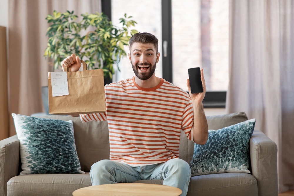 technology, delivery and people concept - smiling man with smartphone and takeaway food in paper bag at home. smiling man using smartphone for food delivery