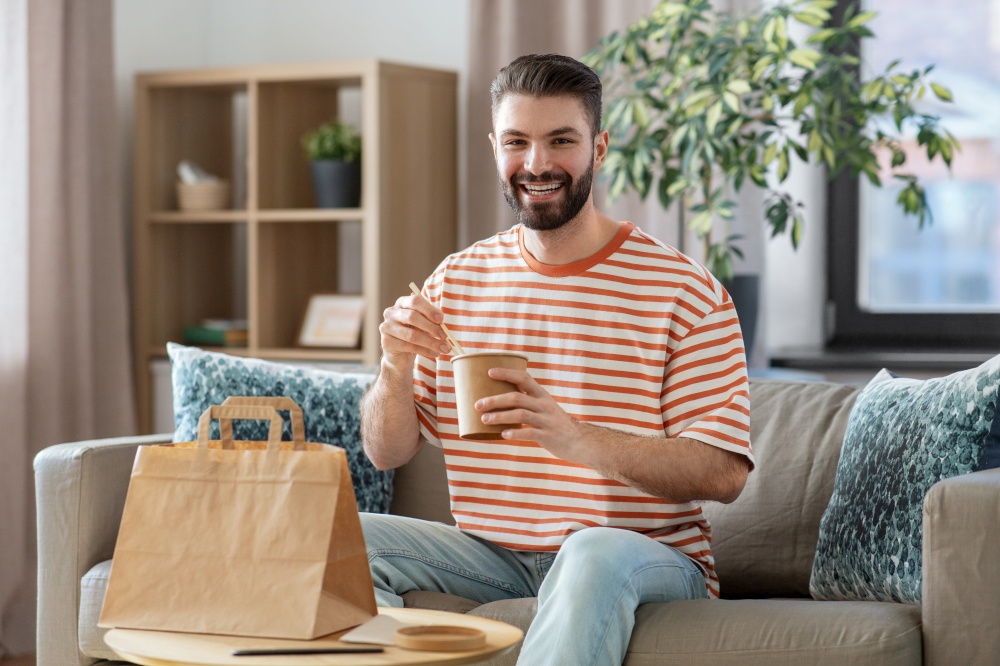 consumption, delivery and people concept - smiling man with chopsticks eating takeaway food at home. smiling man eating takeaway food at home