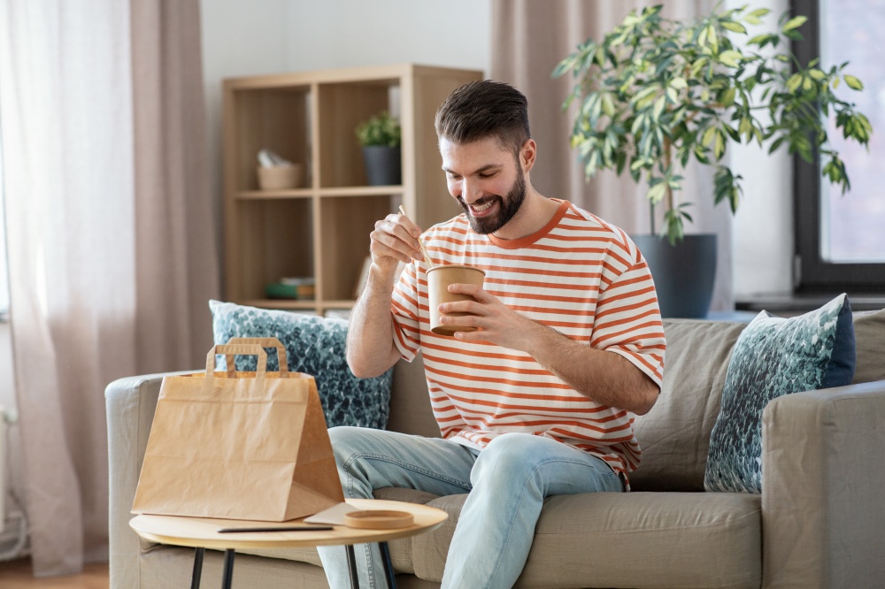 consumption, delivery and people concept - smiling man with chopsticks eating takeaway food at home. smiling man eating takeaway food at home
