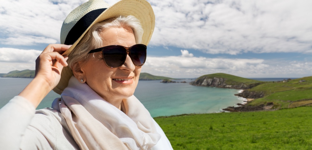 travel, tourism and vacation concept - portrait of happy senior woman in sunglasses and straw hat over atlantic ocean coast in ireland background. happy senior woman in sunglasses and hat on beach