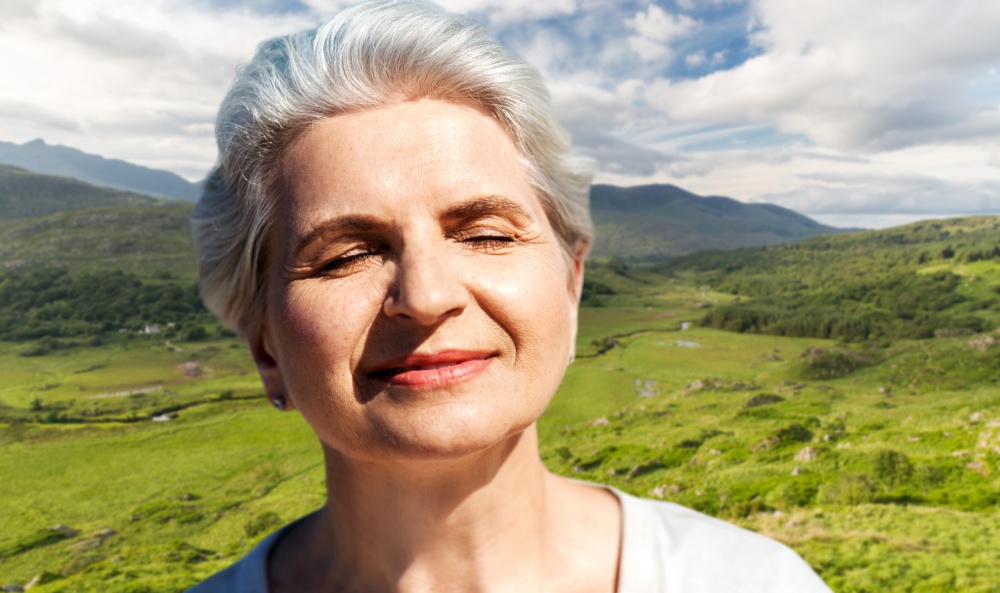 travel, tourism and vacation concept - portrait of happy senior woman enjoying sun over Killarney National Park valley in ireland background. portrait of senior woman enjoying sun on beach