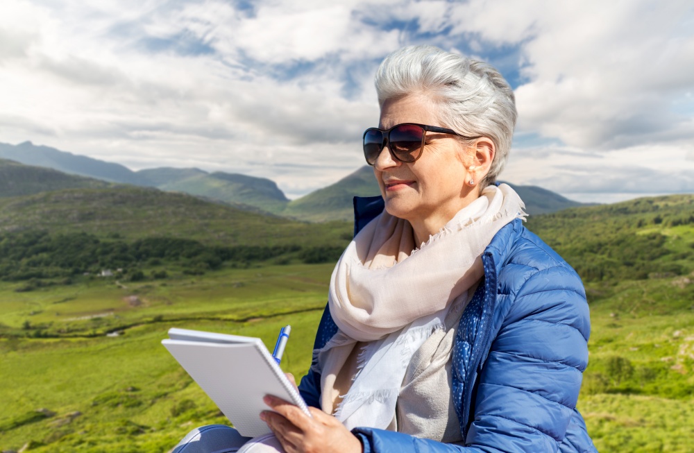travel, tourism and vacation concept - happy senior woman writing to notebook over Killarney National Park valley in ireland background. senior woman writing to notebook on summer beach