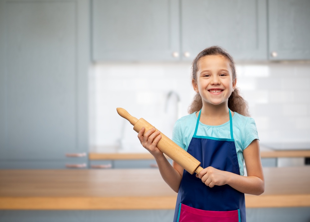 cooking, culinary and baking concept - happy smiling little girl in apron with rolling pin over home kitchen background. smiling little girl in apron with rolling pin
