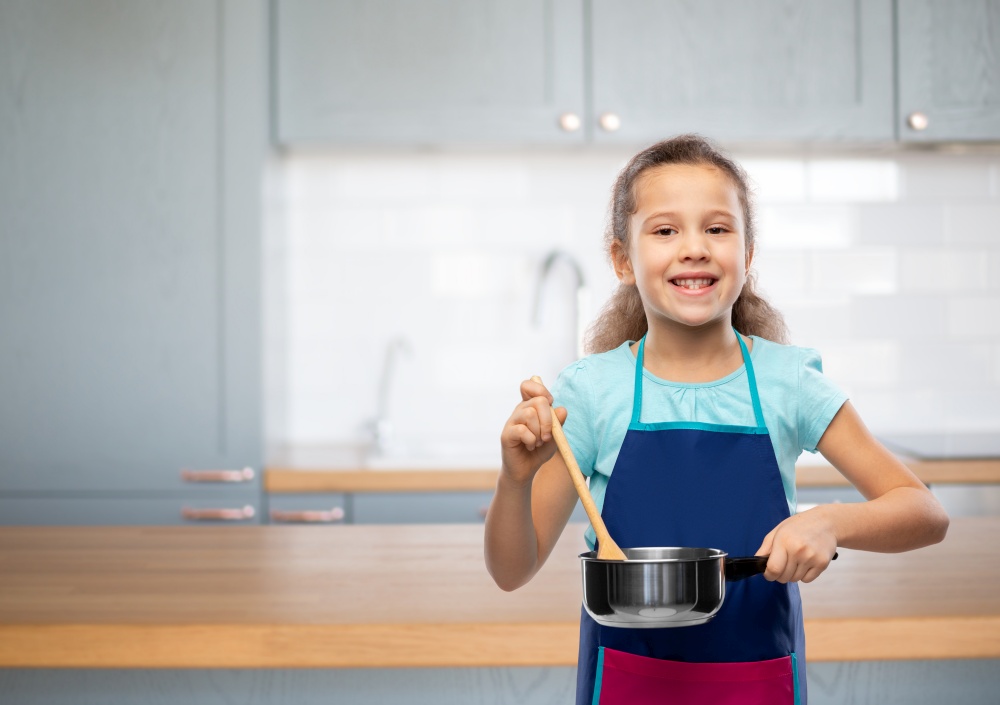 culinary and profession concept - happy smiling little girl in apron with saucepan and wooden spoon cooking food over home kitchen background. little girl in apron with saucepan cooking food