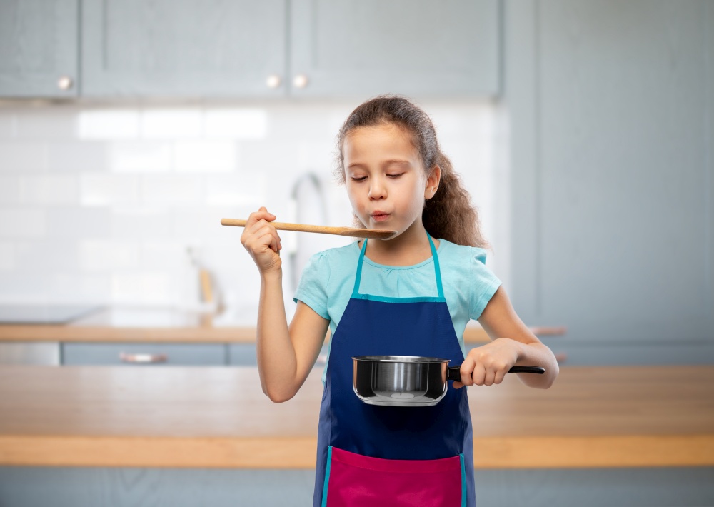 culinary and profession concept - little girl in apron with saucepan and wooden spoon cooking food over home kitchen background. little girl in apron with saucepan cooking food