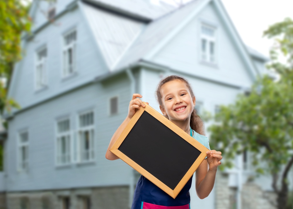 cooking, culinary and profession concept - happy smiling little girl in apron holding chalkboard over living house background. smiling girl in apron with chalkboard over house