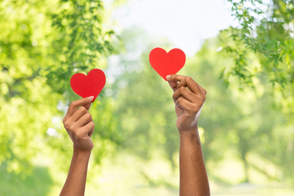 charity, love and health concept - close up of female and male hands holding red heart over green natural background. close up of hands holding red heart