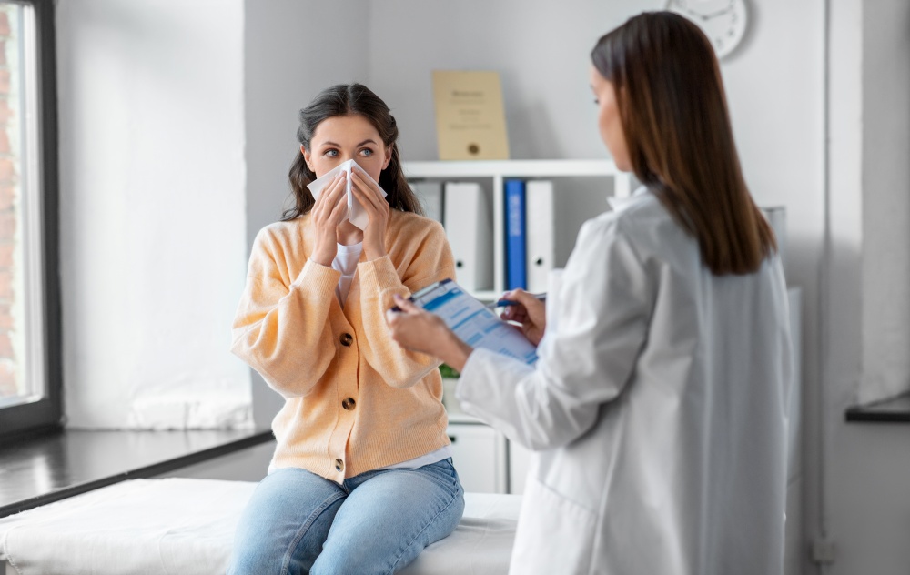 medicine, healthcare and people concept - female doctor with clipboard talking to woman patient blowing her nose with paper tissue at hospital. female doctor and woman blowing nose at hospital