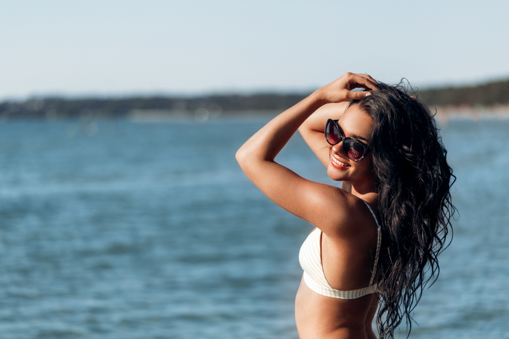 people, summer and swimwear concept - happy smiling young woman in bikini swimsuit and sunglasses posing on beach. smiling young woman in bikini swimsuit on beach