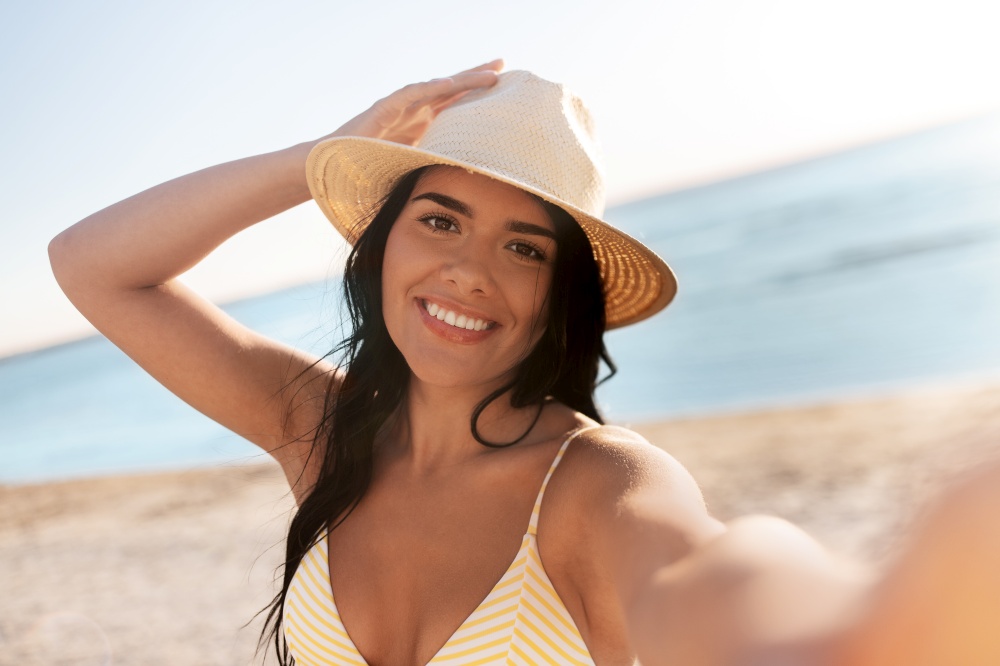 people, summer and swimwear concept - happy smiling young woman in bikini swimsuit and straw hat taking selfie on beach. smiling woman in bikini taking selfie on beach