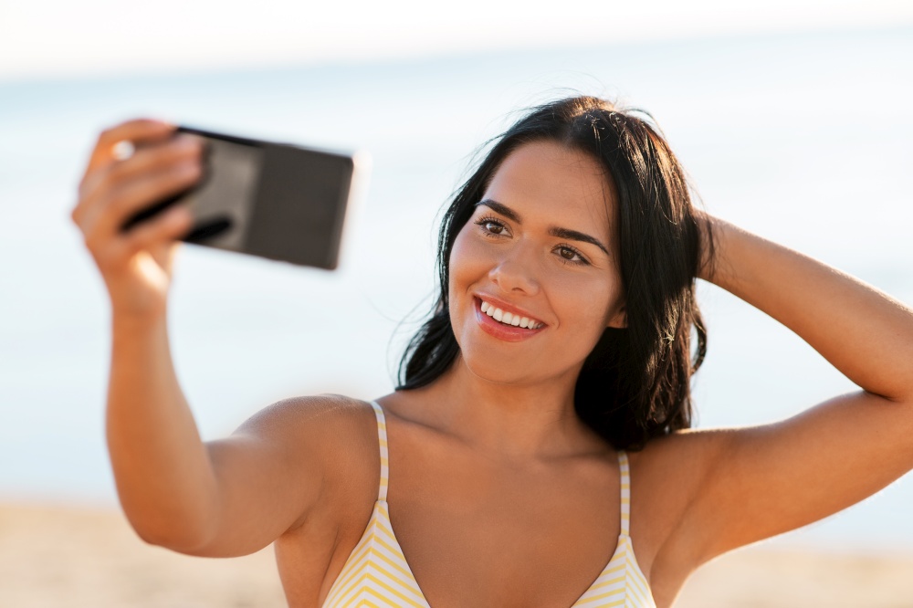 people, summer and swimwear concept - happy smiling young woman in bikini swimsuit taking selfie with smartphone on beach. smiling woman in bikini taking selfie on beach