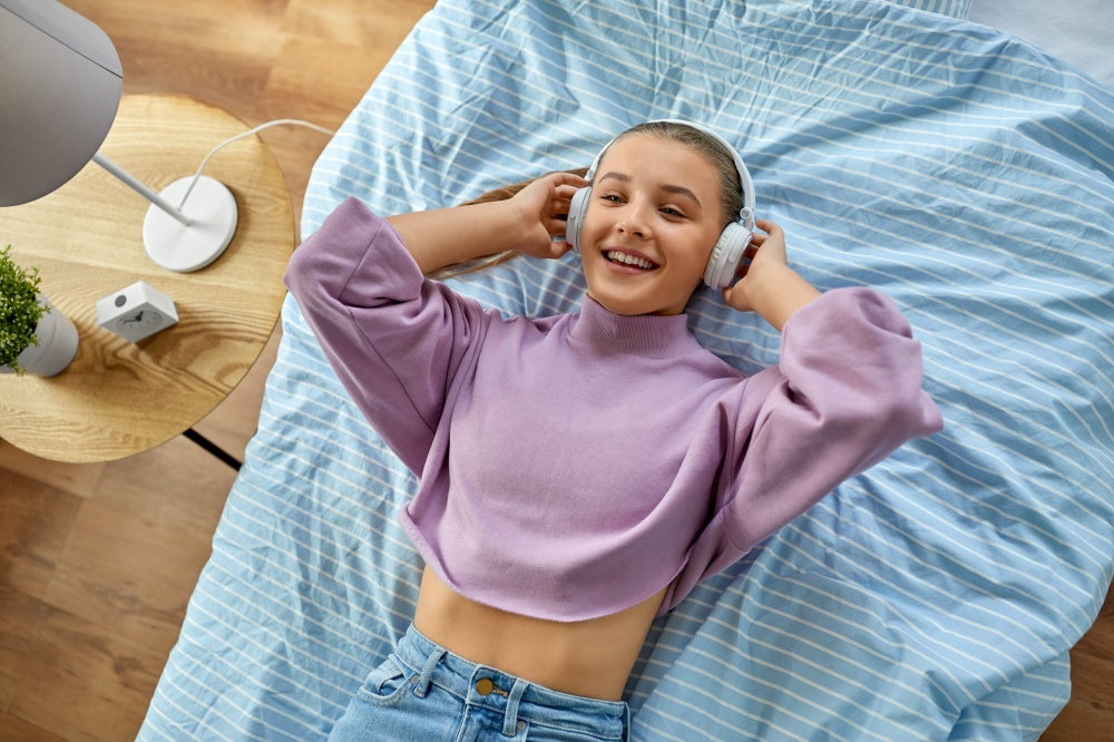 leisure, children and technology concept - girl in headphones lying on bed and listening to music at home. girl in headphones listening to music at home
