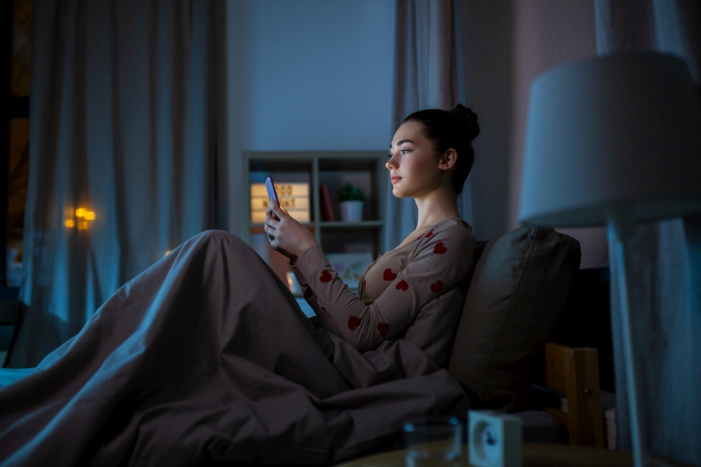 technology, bedtime and rest concept - teenage girl in pajamas with smartphone sitting in bed at night. teenage girl in pajamas with phone in bed at night