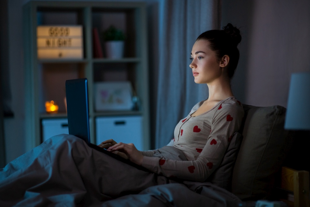 technology, bedtime and people concept - teenage girl with laptop computer sitting in bed at home at night. teenage girl with laptop in bed at home at night