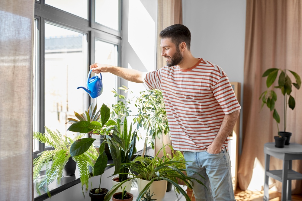 people, nature and plants care concept - happy smiling man watering flowers at home. happy smiling man watering flowers at home