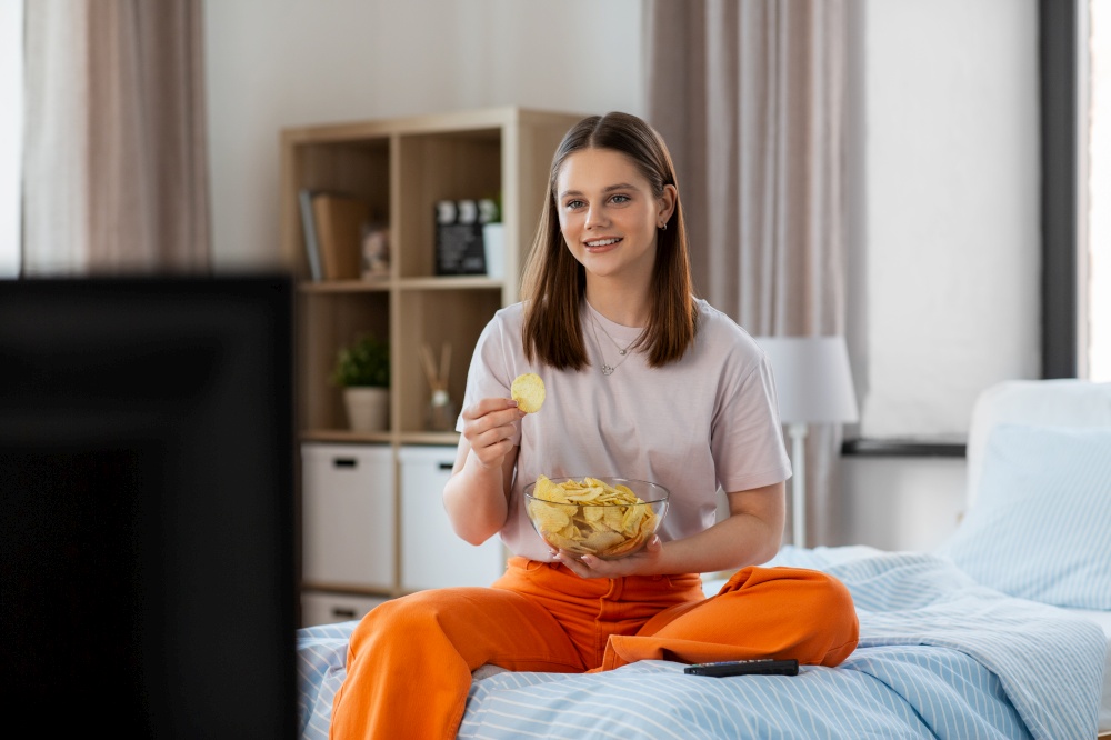 fast food and people concept - happy girl eating crisps and watching tv sitting on bed at home. happy girl eating crisps and watching tv at home