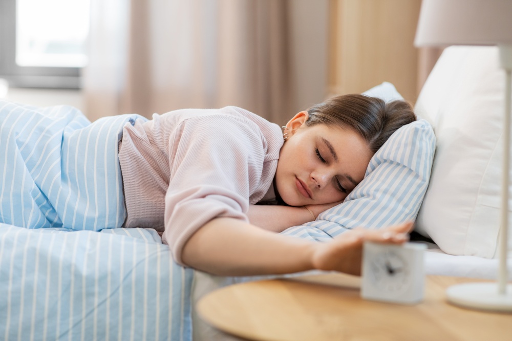 people, bedtime and rest concept - teenage girl sleeping in bed at home in morning and reaching to alarm clock. sleepy girl reaching to alarm clock in morning