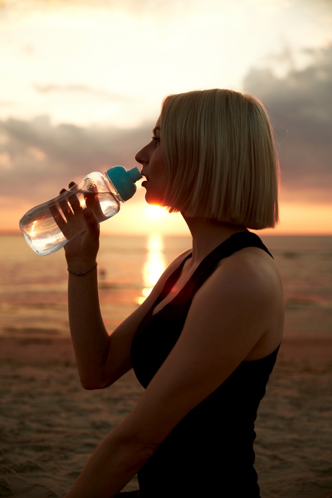 fitness, sport, and healthy lifestyle concept - woman drinking water from bottle on beach over sunset. woman drinking water from bottle on beach