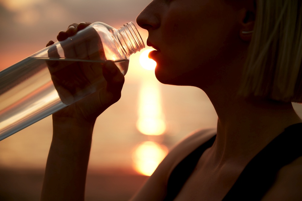fitness, sport, and healthy lifestyle concept - close up of woman drinking water from bottle on beach over sunset. woman drinking water from bottle on beach